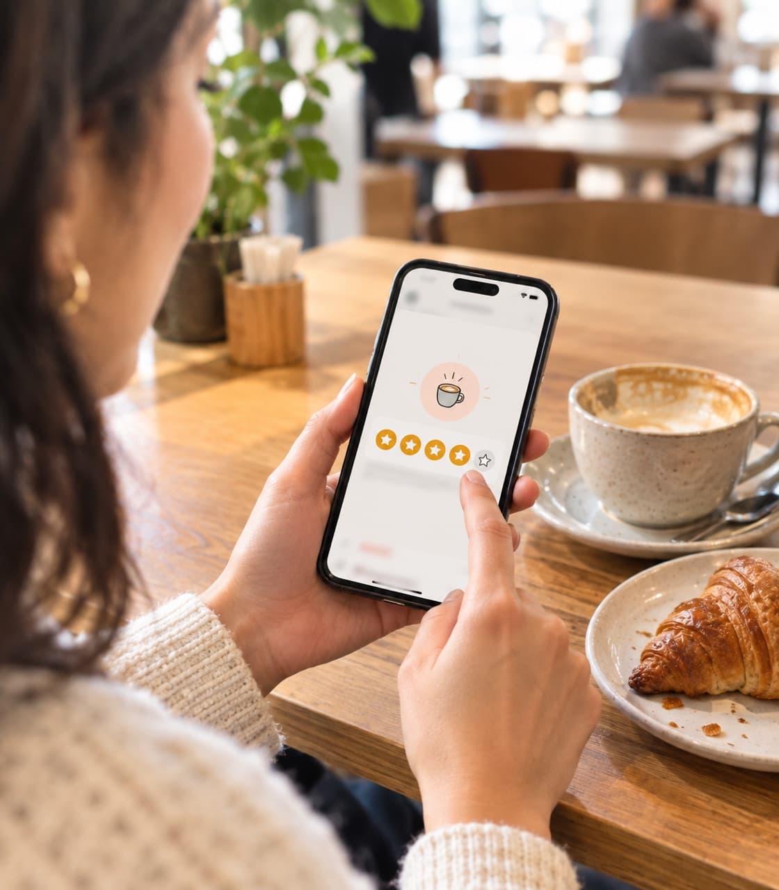 A customer taps a star rating on her phone at a café table, next to a cappuccino and a croissant